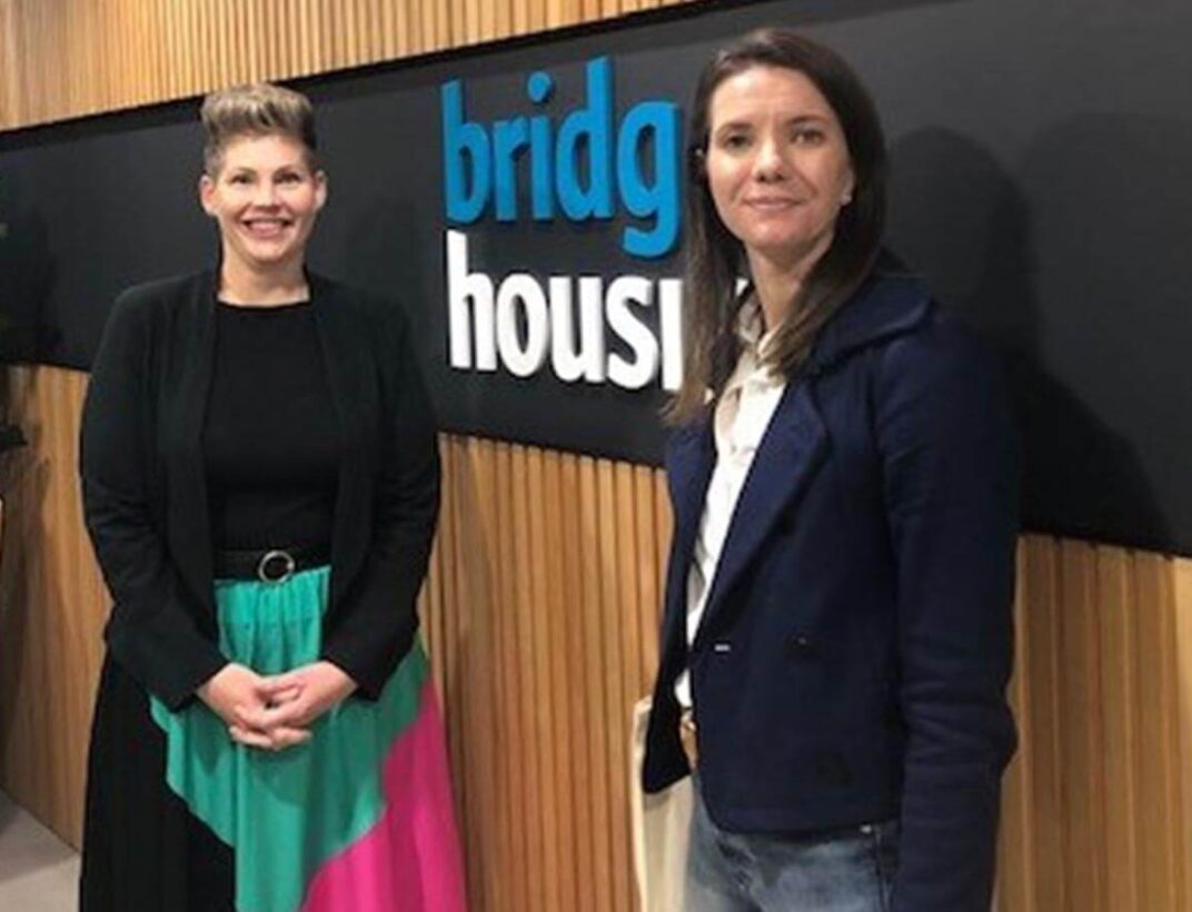 Two women smiling beside "bridge housing" sign