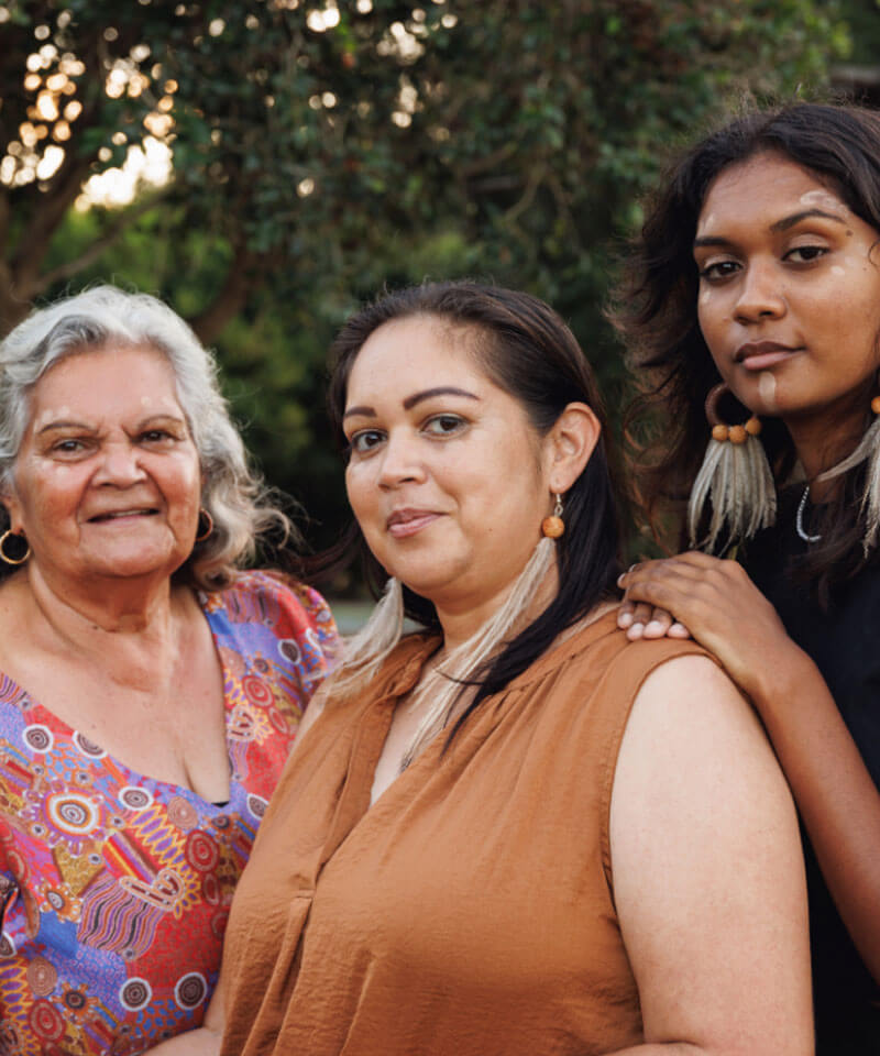 Three aboriginal women posing outdoors, diverse ages, warm expressions.
